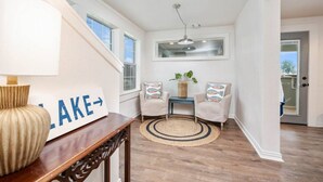 A bright hallway with a wooden floor, a round rug, two armchairs, a small table with a plant, and a sign on the wall.