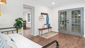 A spacious room with hardwood flooring, a bed, a bench with cushions, a hanging light fixture, and a door with glass panels.