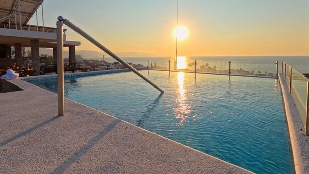 Una piscina con vistas a la puesta de sol sobre el océano, un área de descanso y una zona de estar cubierta.