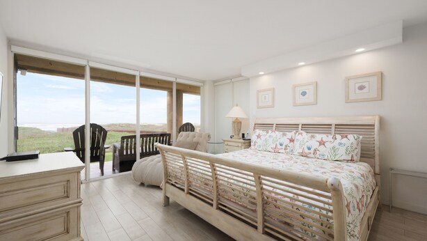 Ein Schlafzimmer mit einem Holzbett, einem Schreibtisch und einem Stuhl. Große Fenster bieten einen Blick ins Freie.