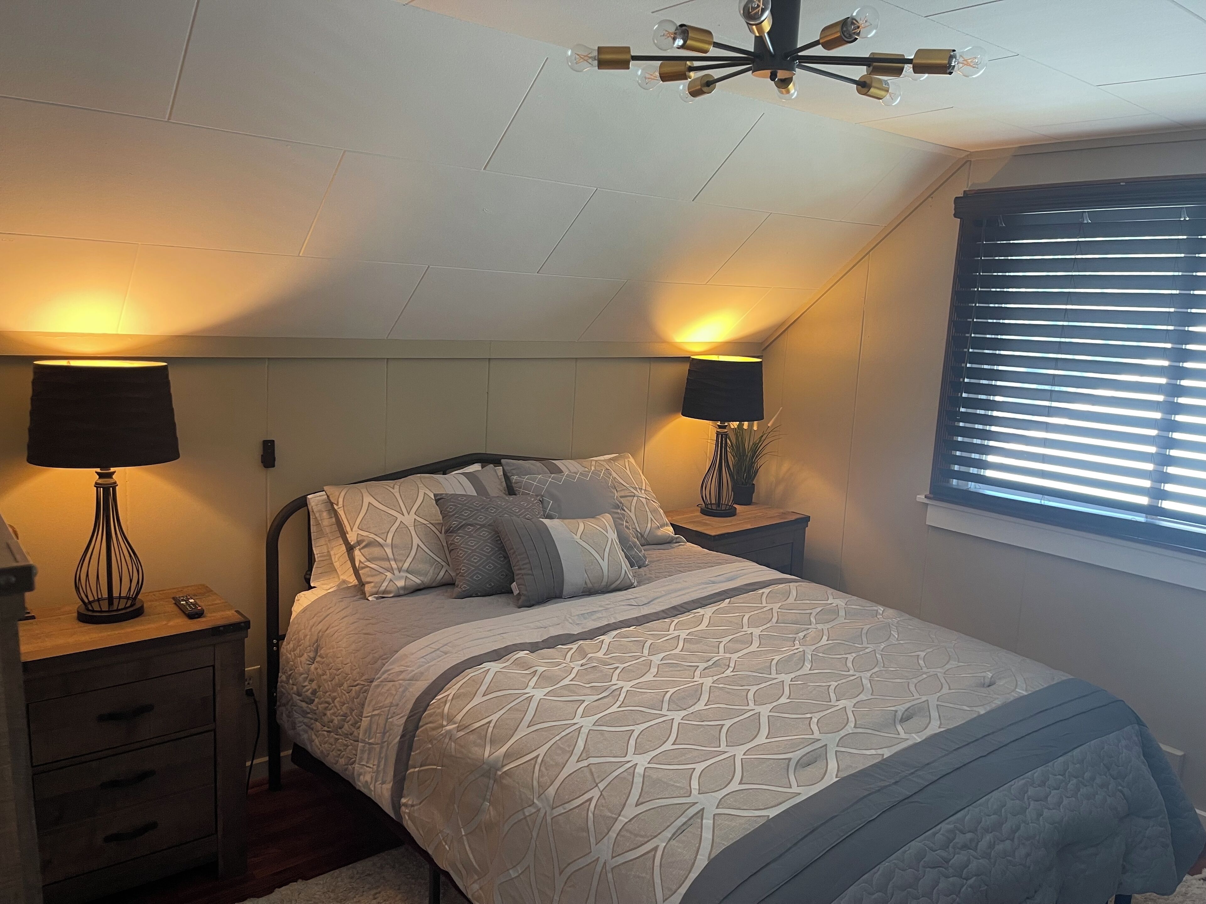 A bedroom with a bed, two bedside tables, lamps, a ceiling light fixture, a window with blinds, and a wall-mounted plant.