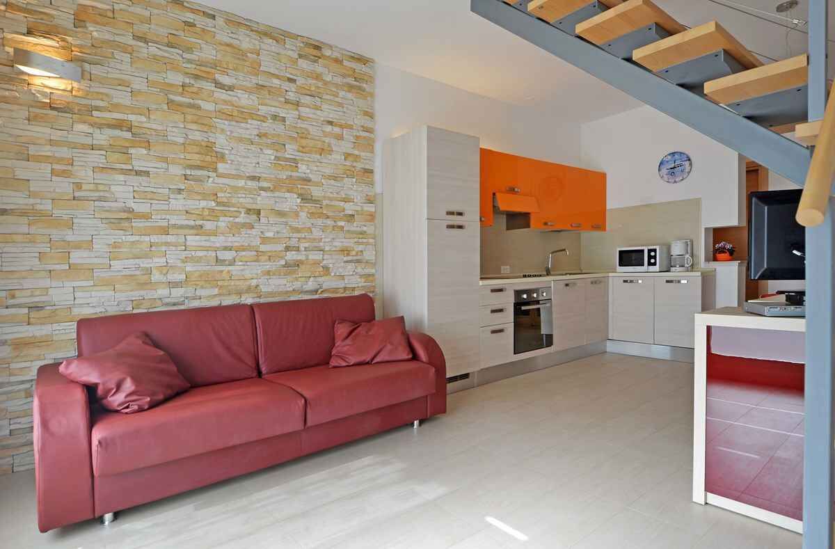 Una sala de estar moderna con un sofá rojo, una pared de piedra y una cocina con armarios blancos y encimera naranja.