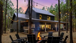 Une cabane avec un foyer entouré de fauteuils Adirondack, située dans une forêt et illuminée par des guirlandes lumineuses suspendues au-dessus.