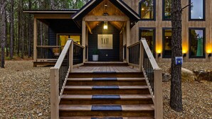 Une cabane en bois avec un escalier menant à une entrée couverte, entourée d’arbres et de gravier.