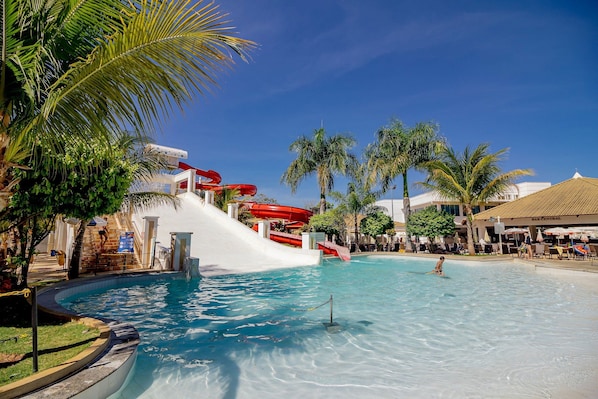 Un parque acuático con una piscina grande, un tobogán de agua rojo, palmeras y un edificio con la inscripción 'BAR CENTRAL'.