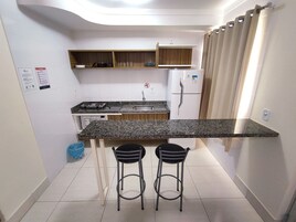 Cocina compacta con encimera de granito, dos taburetes de bar, un refrigerador y armarios de madera.