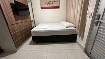 Uma cama pequena e bem arrumada em quarto com parede de painel de madeira, televisão fixada e um quadro na parede.