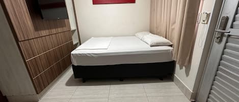 A small, neatly made bed in a room with a wooden panel wall, a mounted TV, and a painting on the wall.