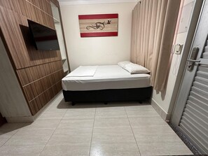 Un petit lit, bien fait, dans une pièce dont un mur est recouvert de panneaux de bois, avec un téléviseur fixé au mur et un tableau accroché.