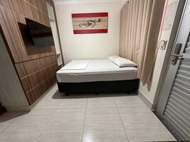 Una cama pequeña y ordenada en una habitación con pared de paneles de madera, televisor montado y un cuadro en la pared.