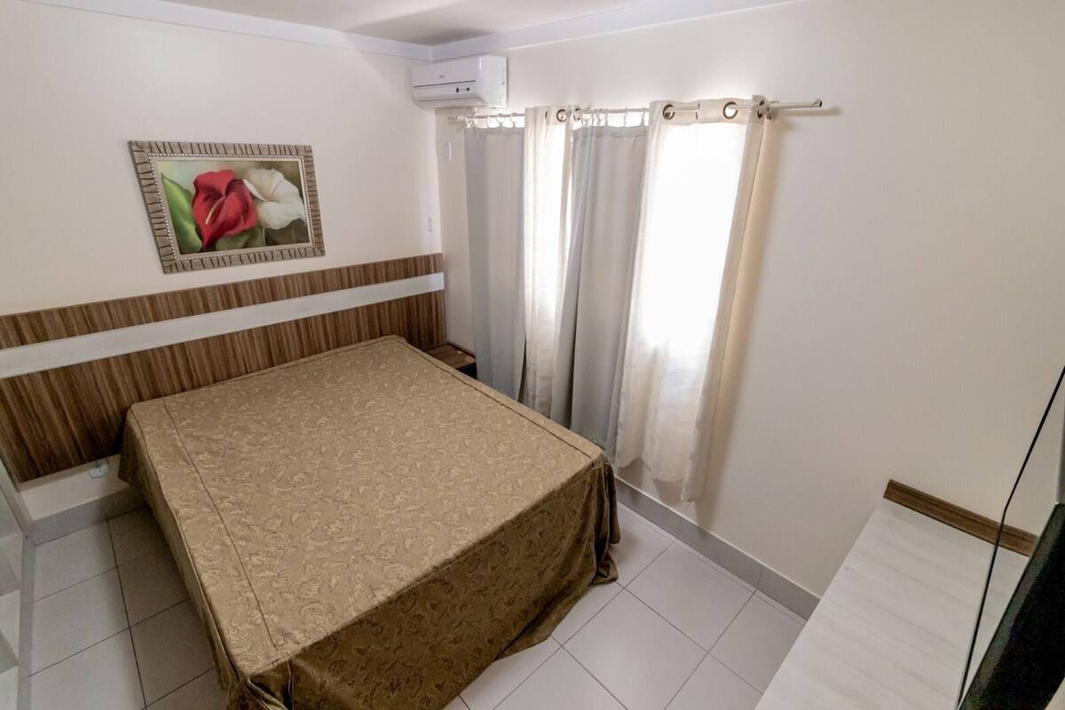A bedroom with a bed, a wall-mounted air conditioner, a window with curtains, and a framed picture on the wall.