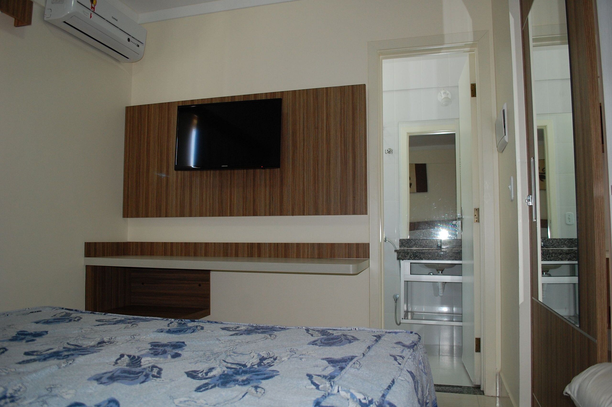 Un dormitorio con cama, televisor montado en una pared de paneles de madera y baño visible a través de una puerta abierta.