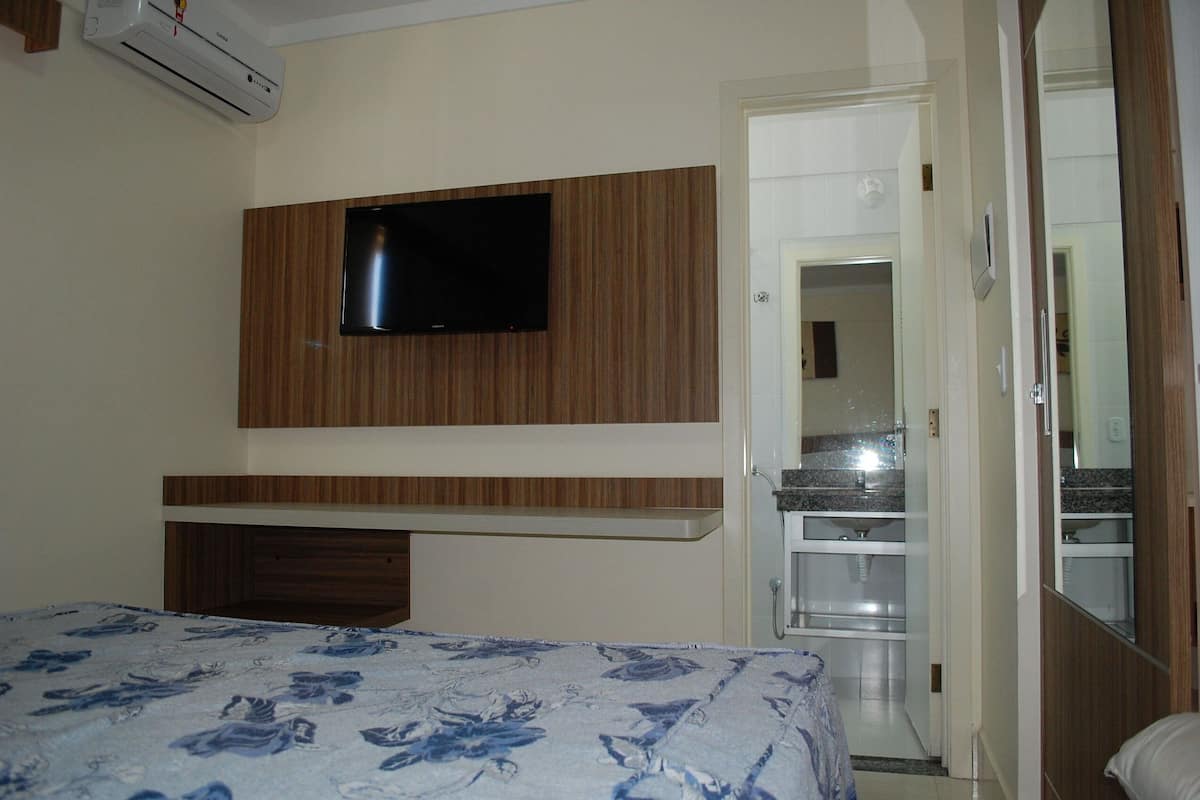 A bedroom with a bed, a TV mounted on a wooden panel wall, and a bathroom visible through an open door.