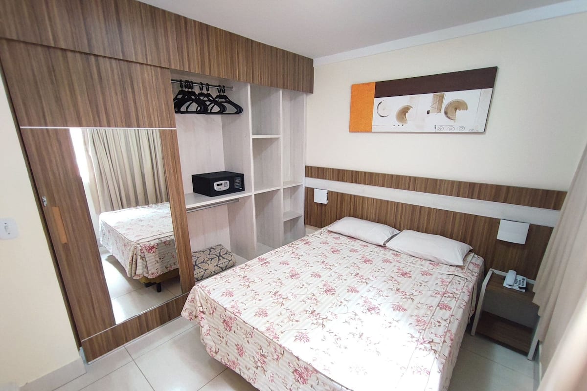 A bedroom with a bed, a wardrobe, a bedside table, and a wall-mounted shelf.