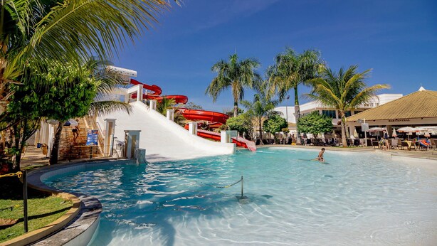A water park with a large pool, a red water slide, palm trees, and a building labeled "BAR CENTRAL". - LACQUA II (Caldas Novas)
