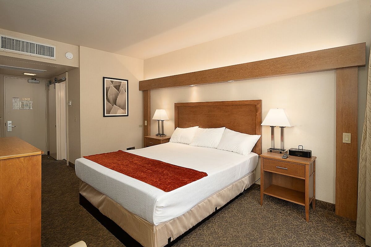 A hotel room with a large bed, two bedside tables with lamps, a wooden headboard, a red throw blanket, and a framed picture on the wall.