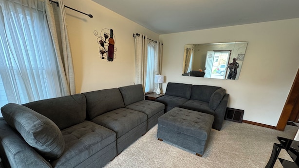 Ein Wohnzimmer mit einem grauen Ecksofa, einem passenden Hocker, einem Beistelltisch mit Lampe und einem Spiegel an der Wand.