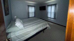 Ein Schlafzimmer mit einem Bett, zwei Fenstern mit Vorhängen, einem Deckenventilator und einem Flachbildfernseher an der Wand.