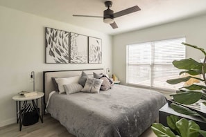A bedroom with a bed, a ceiling fan, a window with blinds, and a plant.