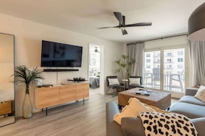 A modern living room with a flat-screen TV, a ceiling fan, a sofa, a coffee table, and a view of the outside balcony.