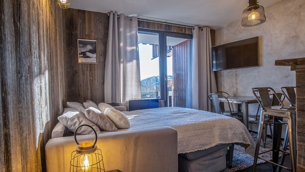 A bedroom with a bed, a bedside table with a lantern, a TV mounted on the wall, and a view of the outdoors. - Appartement du Rif - Welkeys (Huez)