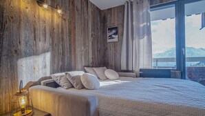 A bedroom with a large bed, wooden walls, a nightstand with a lamp, and a view of mountains through a large window. - Appartement du Rif - Welkeys (Huez)