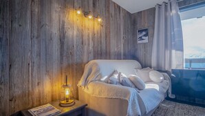 A cozy room with a wooden wall, a bed with a white blanket, a bedside table with a lamp, and a window with a view of mountains. - Appartement du Rif - Welkeys (Huez)