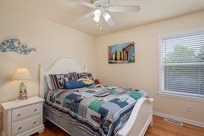 A bedroom with a bed, nightstand, lamp, ceiling fan, artwork, and a window with blinds.