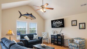 Una sala de estar con ventilador de techo, una gran decoración de pez, un televisor que muestra 'Coastal Carolina Vacations and Sales' y vistas del exterior a través de la ventana.