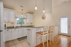 Une cuisine avec des meubles blancs, un îlot central en bois avec des chaises, une fenêtre donnant sur un beau paysage et une horloge au mur.