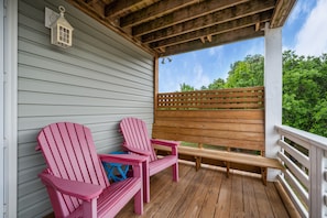 A wooden deck with two Adirondack chairs and a bench, a white railing, and a view of greenery.