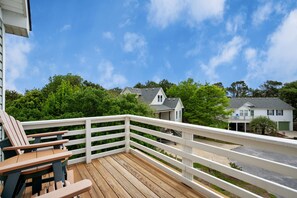 Une terrasse en bois avec des rampes blanches, un espace salon avec des chaises, et une vue sur des maisons et des arbres en arrière-plan.