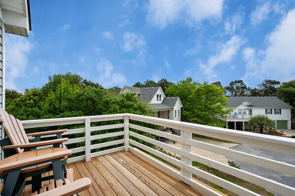 Une terrasse en bois avec des rampes blanches, un espace salon avec des chaises, et une vue sur des maisons et des arbres en arrière-plan.