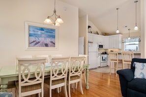A modern kitchen with white cabinets, a dining area with a glass table, and a painting of a beach scene.