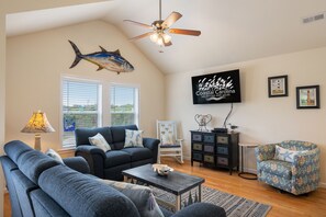 A living room with a ceiling fan, a large fish decoration, a TV displaying 'Coastal Carolina Vacations and Sales', and a view of the outdoors through the window.