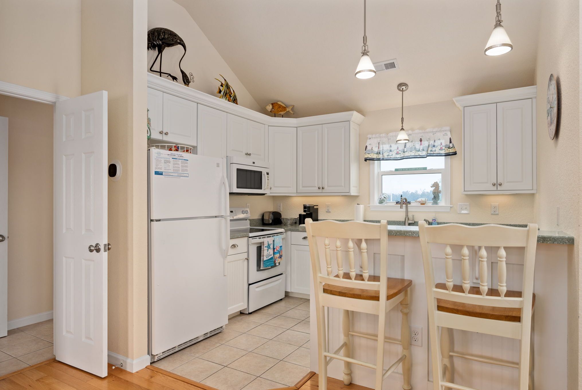 A kitchen with white cabinets, a microwave, an oven, a refrigerator, two wooden chairs, and pendant lights.