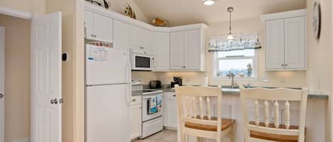 A kitchen with white cabinets, a microwave, an oven, a refrigerator, two wooden chairs, and pendant lights.