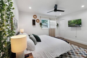 A bedroom with a bed, a ceiling fan, a window with curtains, a TV on the wall, and a plant.