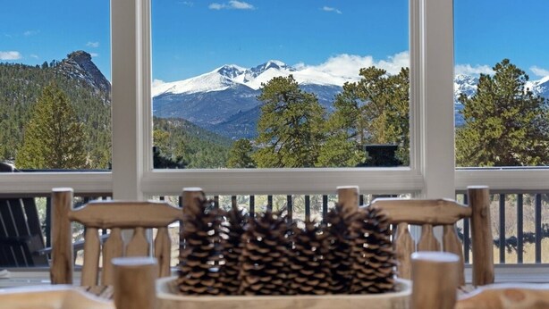 A view from a window overlooking snow-capped mountains, a wooden deck, and pine cones. - Spectacular Views of Long's Peak- 10 min to town-3 full bath plus 2 half bath (Estes Park)