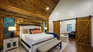 A bedroom with a wooden ceiling, a large bed with multiple pillows, a bench, and a sliding barn door. - It's the Little Things, Creekside, Hot Tub, Firepit, Great Location (Broken Bow)