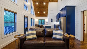 A cozy living room with a brown leather couch, wooden floors, and a kitchen in the background. - It's the Little Things, Creekside, Hot Tub, Firepit, Great Location (Broken Bow)