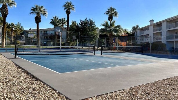 An outdoor tennis court with a net, surrounded by a fence and palm trees.