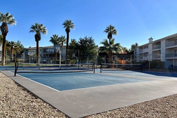 An outdoor tennis court with a net, surrounded by a fence and palm trees.