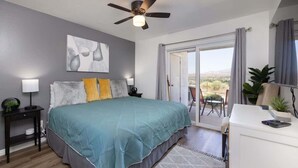 A bedroom with a bed, a ceiling fan, a nightstand, a desk, a chair, and a view of the outdoors.