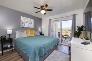 A bedroom with a bed, a ceiling fan, a nightstand, a desk, a chair, and a view of the outdoors.
