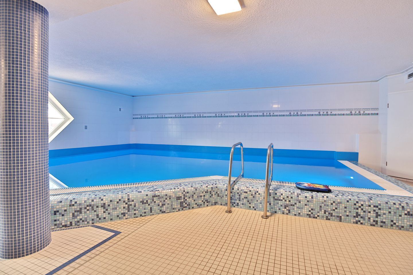 An indoor swimming pool with a blue and white tiled floor, a pool ladder, and a decorative wall.