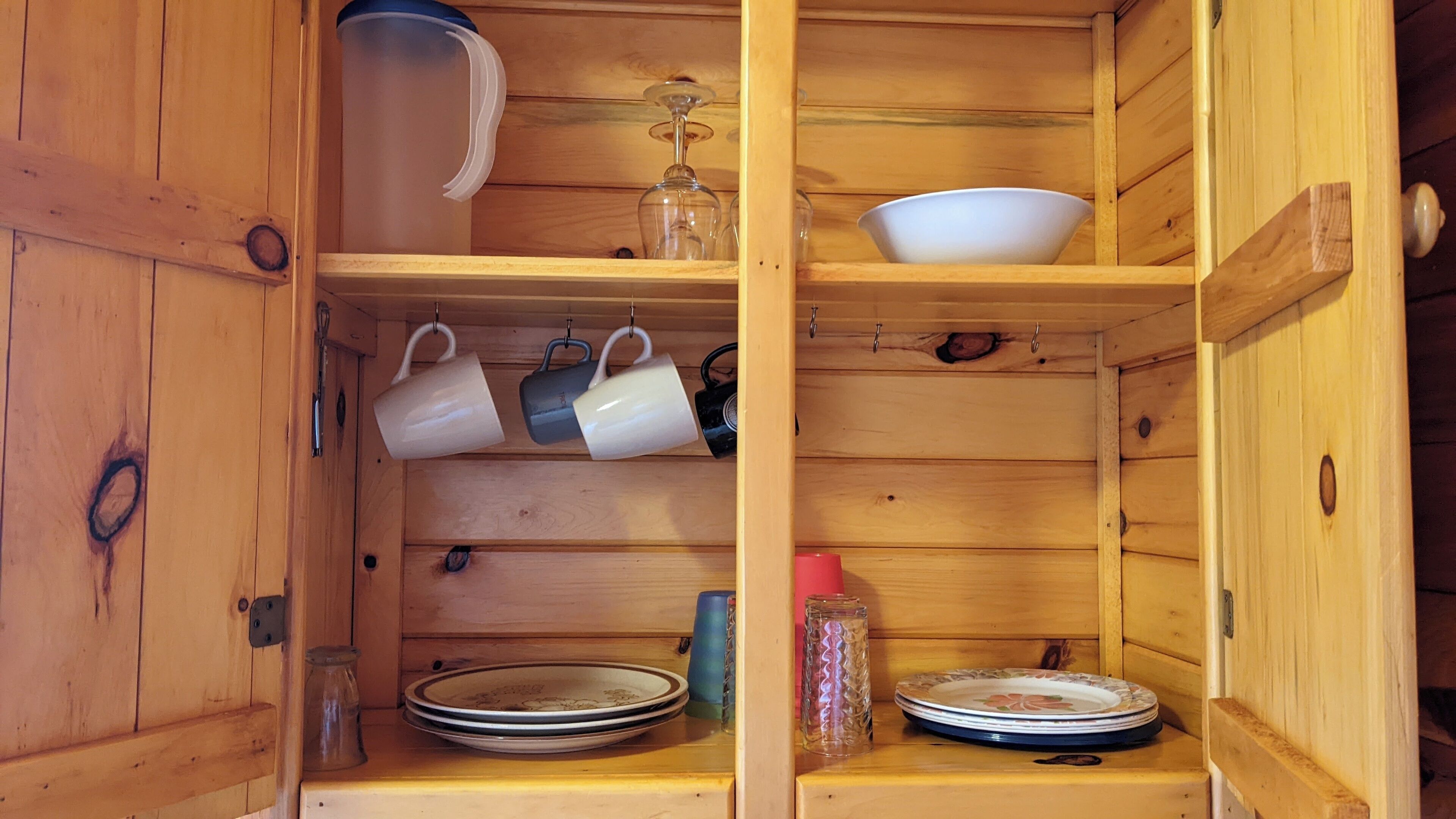 Un armario de madera con estantes que guardan diversos utensilios de cocina, incluyendo platos, tazas y un tarro de vidrio.