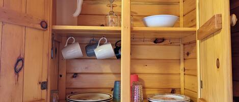 Un armario de madera con estantes que guardan diversos utensilios de cocina, incluyendo platos, tazas y un tarro de vidrio.