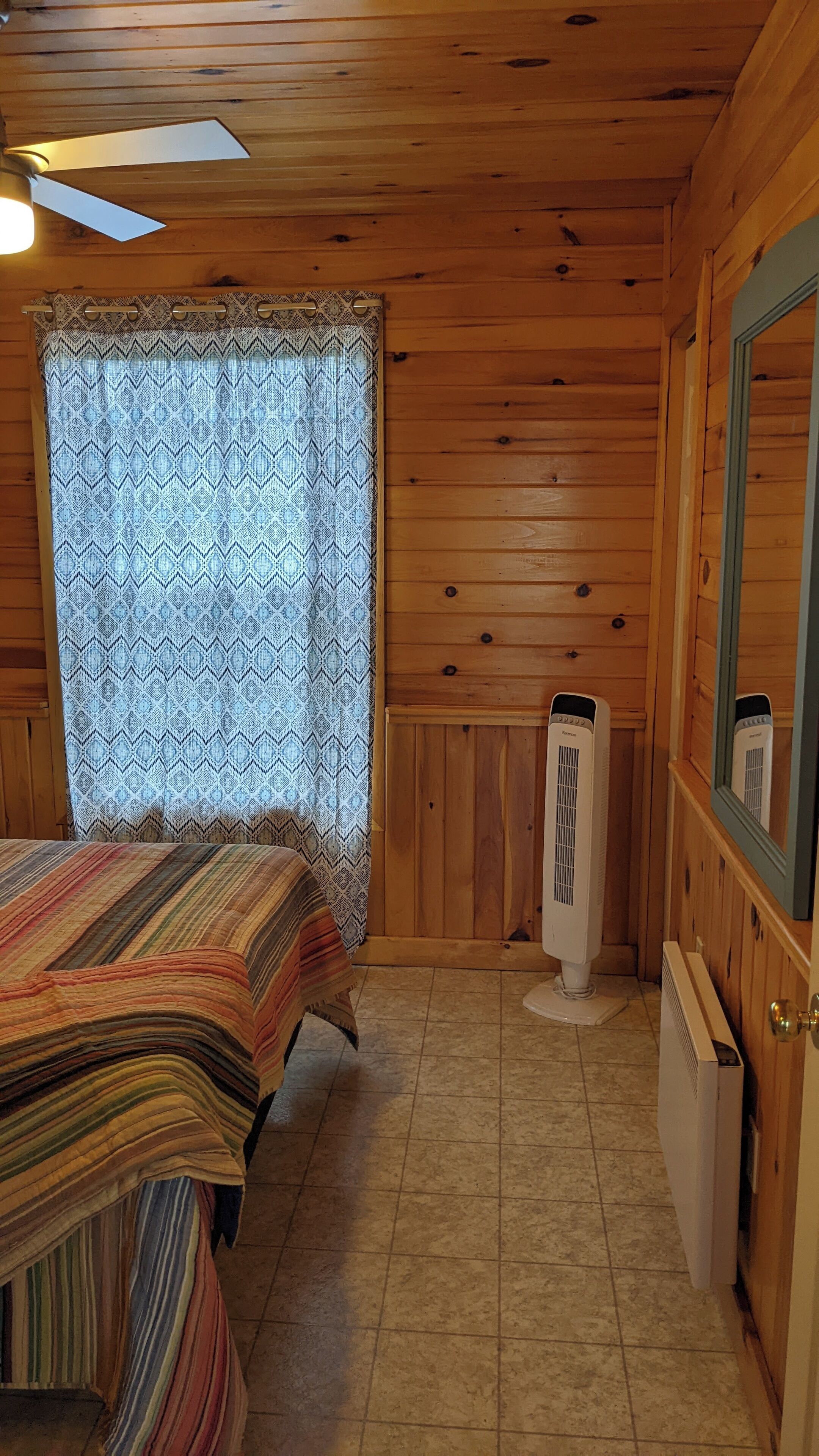 Camera in baita di legno con un letto, una ventola a soffitto, una finestra con tende a motivi e un termosifone a parete.