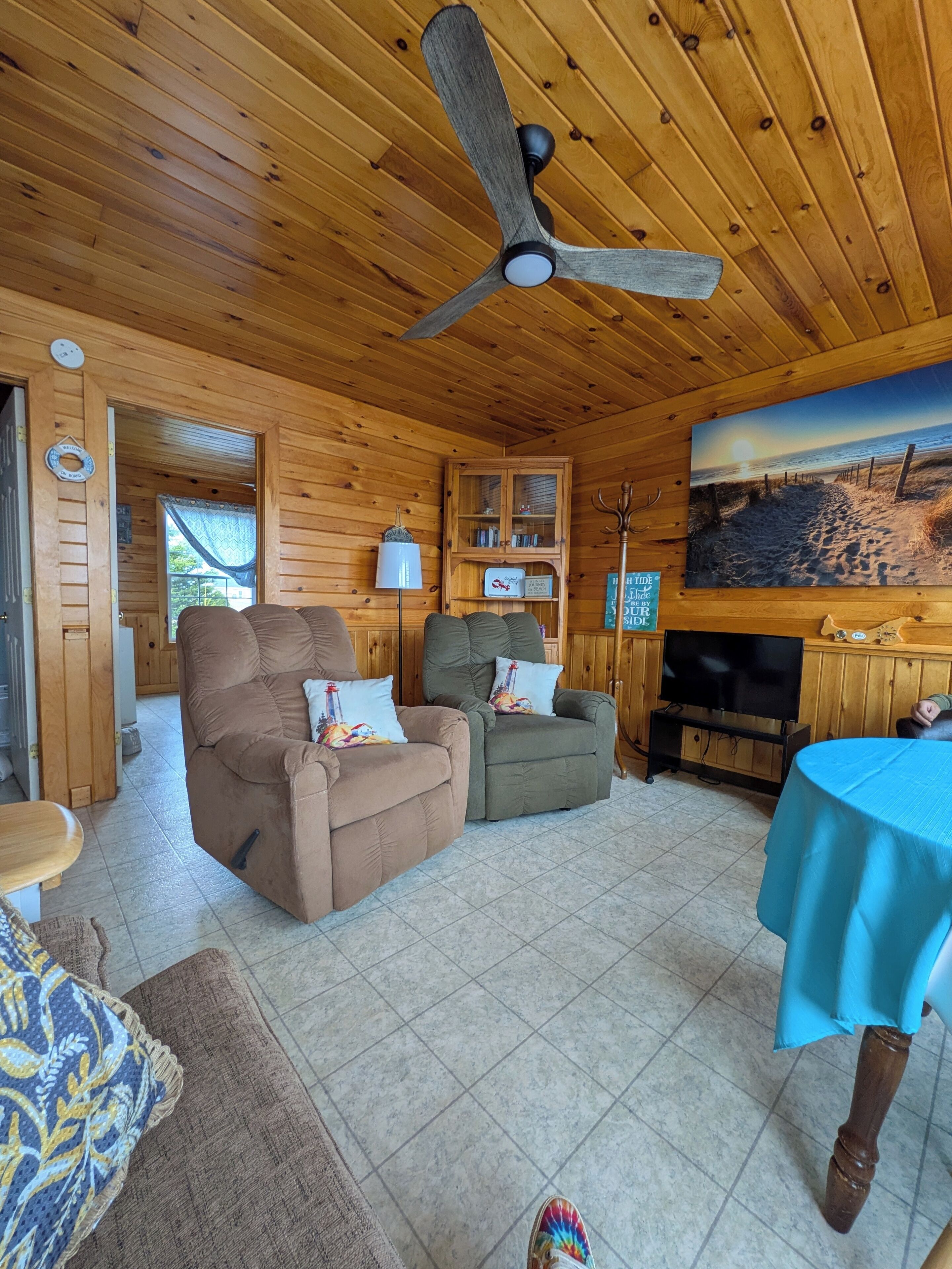 Interior acollidor d'una cabana amb sostre de fusta, ventilador de sostre, una llar de foc, televisió i un quadre de platja a la paret.